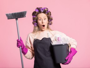 The surprised shocked woman is holding brush to clean floor and mop bucket with cleaning fluid. The girl saw huge mess is horrified by the amount of dust. Housewife concept on pink studio background