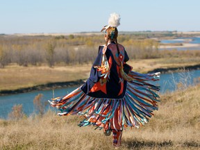 Back of person in First Nations shawl performing a Fancy Shawl Dance in a grass field with a river in the background