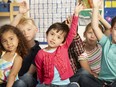 Group of elementary age schoolchildren raising hands to answer question in classroom.