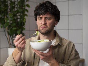 A man in a beige shirt eats a tasteless salad in the kitchen.