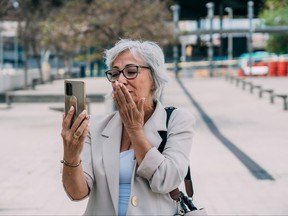Elderly grey-haired female in glasses sends an air kiss while chatting on video call via cellphone
