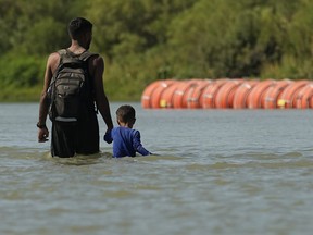 FILE - Migrants walk past large buoys being used as a floating border barrier on the Rio Grande, Aug. 1, 2023, in Eagle Pass, Texas. Texas attorneys asked federal appeals court judges Thursday, Oct. 5, 2023 to let the state keep in place a floating barrier of large concrete-anchored buoys to block migrants from crossing the Rio Grande -- a barrier the Biden administration says was illegally deployed without needed federal authorization.