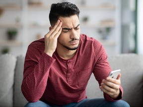 Young man sitting on sofa at home, appearing confused while looking at smartphone.