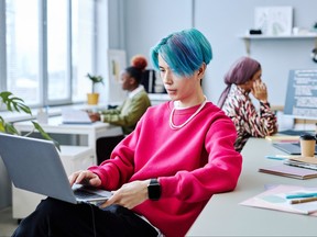 Young man with blue hair using laptop in modern office with two co-workers behind him.