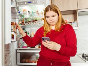 Portrait of pensive woman standing near open refrigerator, holding mobile phone