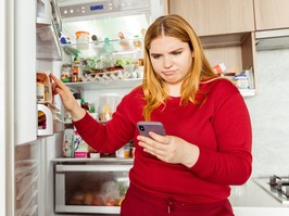 Portrait of pensive woman standing near open refrigerator, holding mobile phone