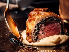 Closeup of hand holding a spoon next to a dish with beef wellington and mashed potatoes.