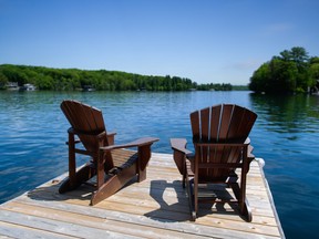Backs of two Adirondack chairs sitting on a wooden pier facing the calm water of a lake in Muskoka.
