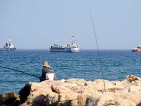 A fisherman sits on a rock as the Open Arms ship and another vessel sail next to the ship Jenifer, of the World Central Kitchen carrying food aid for the Gaza Strip, as they prepare to set sail close to the port of Larnaca in Cyprus on March 30, 2024, amid the ongoing conflict between Israel and the Palestinian Hamas group.