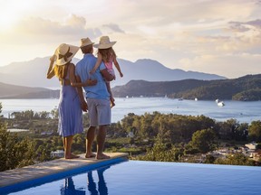A family on summer holidays stands by the swimming pool and enjoys the beautiful sunset behind the mountains and the sea