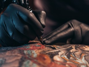 Hands of a tattoo artist wearing black gloves and holding a machine while tattooing a mans back.
