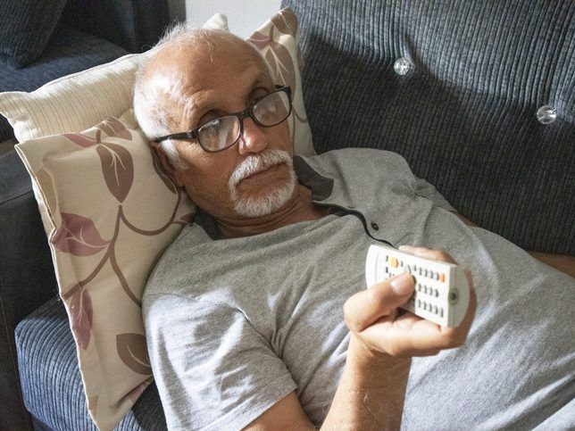 Senior man laying down on the sofa, watching tv and holding the remote control.