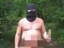 Naked man (private area blurred out) wearing balaclava and clogs while holding stick in woods near castle in Slovakia.