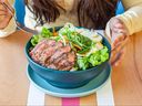 Woman eating a mixed green salad with sliced steak served in a big bowl.
