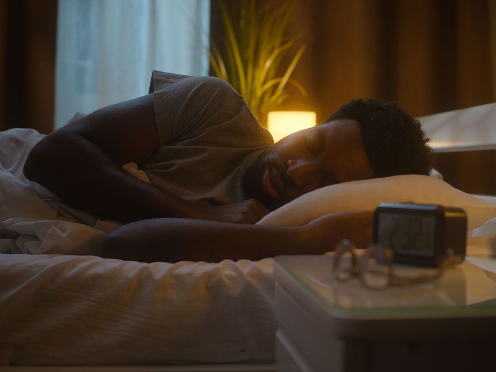 Irritated disturbed ethnic African American man male guy sleeping lying down bed napping waking awake insomnia check time clock digital alarm insomniac sleepless late night at home illuminated bedroom