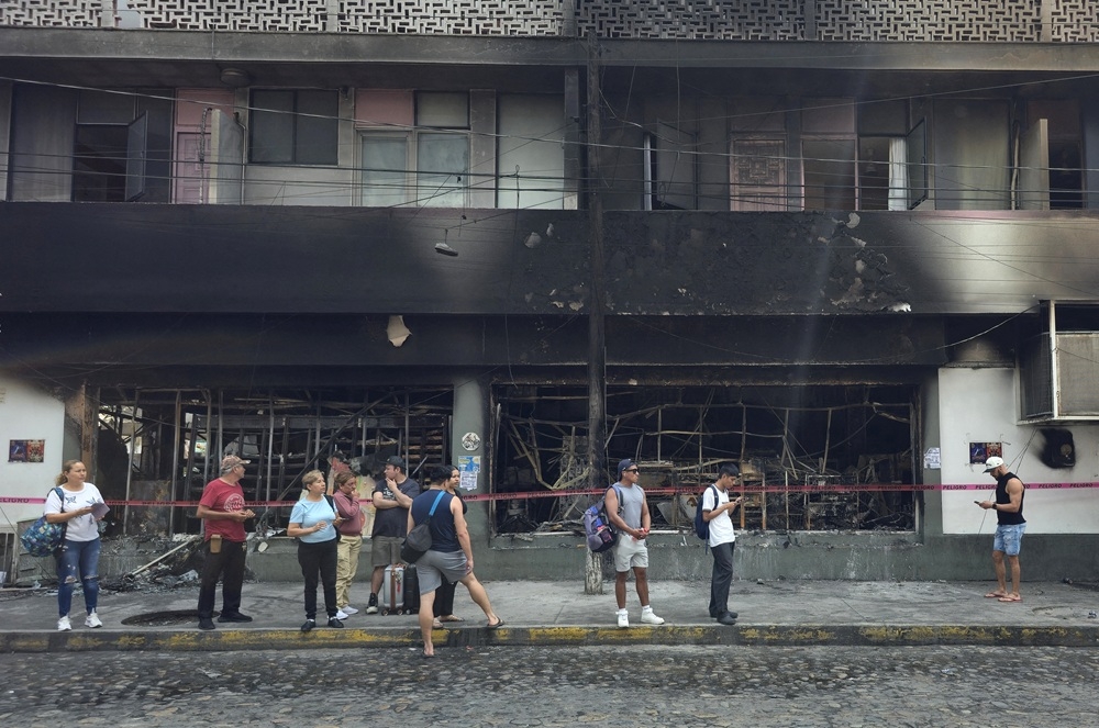  People stand next to a burned building in Puerto Vallarta, Jalisco state, Mexico, on Feb. 23, 2026. Mexico has deployed 10,000 troops to quell clashes sparked by the killing of the country’s most wanted drug lord, which have left dozens dead, officials said on February 23. Nemesio “El Mencho” Oseguera, leader of the Jalisco New Generation Cartel (CJNG), was wounded on February 22 in a shootout with soldiers in the town of Tapalpa in Jalisco state and died while being flown to Mexico City, the army said. ARTURO MONTERO/GETTY IMAGES