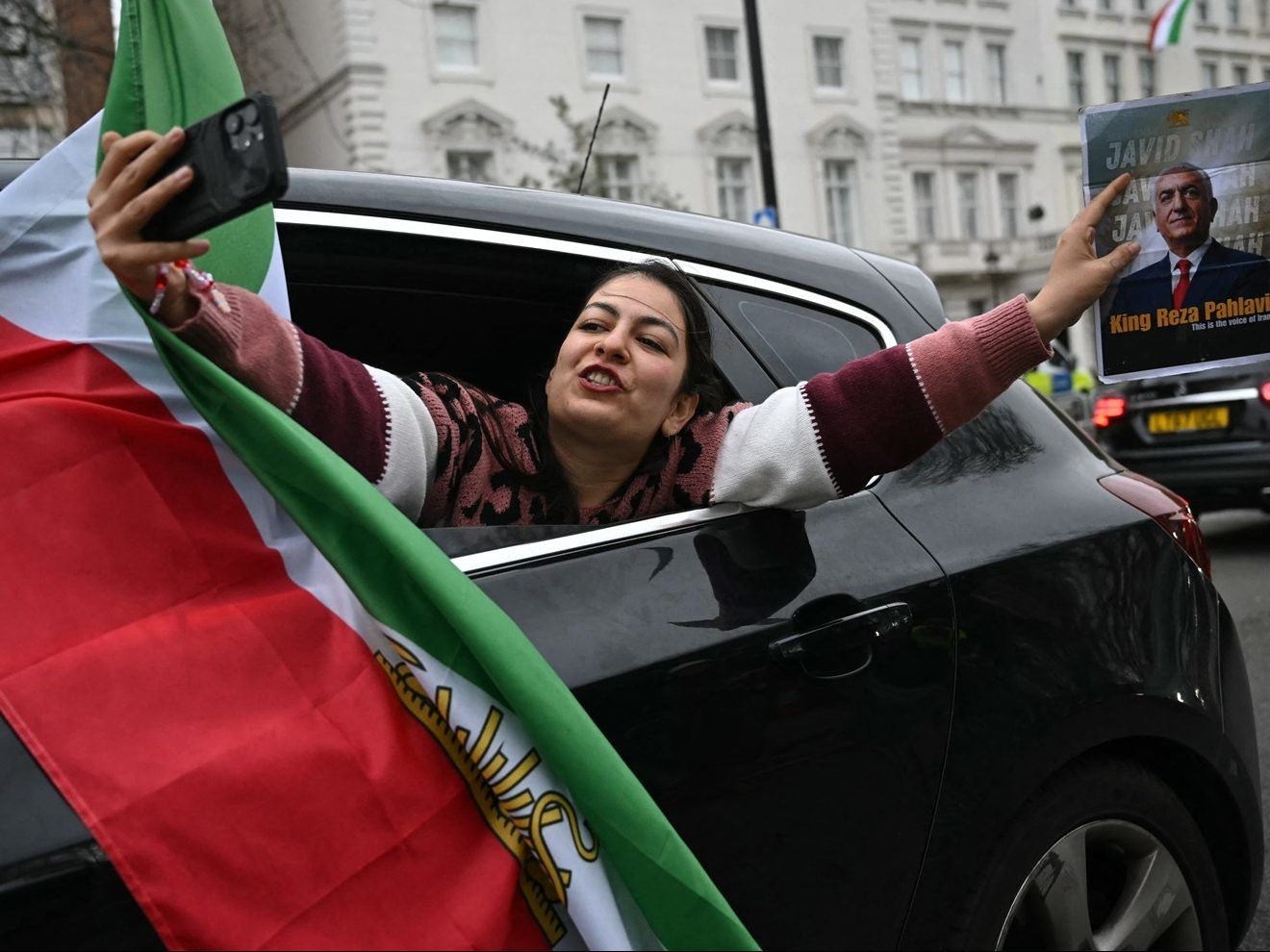 A woman takes a selfie photo while holding a photo of Iranian opposition figure and son of the last Shah of Iran, Reza Pahlav during a drive-by celebration outside the Iranian Embassy in central London, on March 1, 2026.