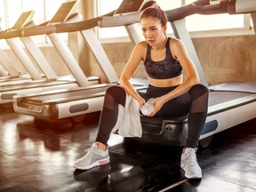 Beautiful Asian young woman tired taking a break from running or exercise sitting on treadmill machine drinking water and towel sweat in fitness gym healthy .girl in sportswear workout rest in morning