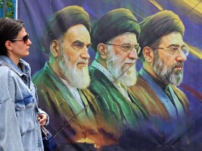 A woman walks past a banner featuring Iran's late supreme leaders Ayatollah Ruhollah Khomeini (left) and Ayatollah Ali Khamenei (centre) next to newly elected supreme leader Ayatollah Mojtaba Khamenei, in Tehran on April 19, 2026.