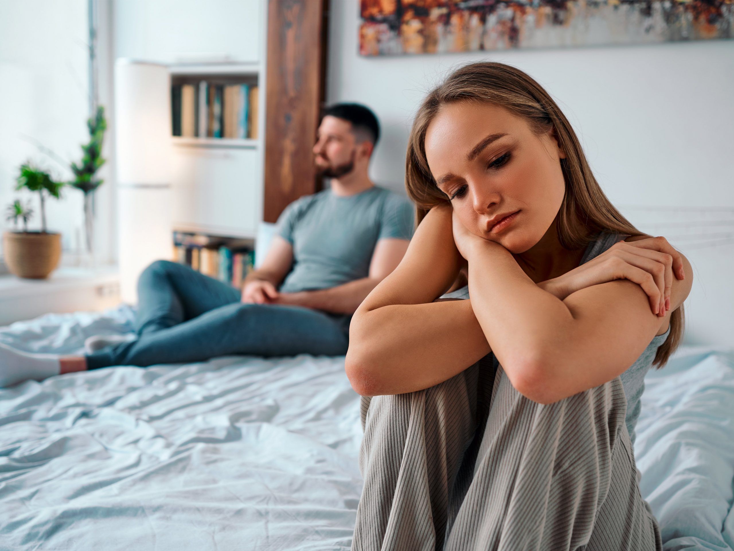 Upset young woman in home clothes sitting on the bed, in the background sitting man. Family troubles.