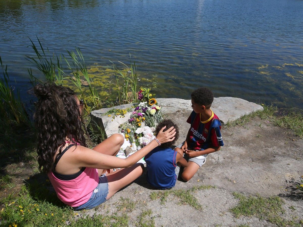 Teryn Allred and her two sons Taevis, 4, and Jikai, 8, sit next to flowers and other mementoes at a small pond in the Legacy neighbourhood in southeast Calgary on July 13, 2026. A day earlier, a child drowned near that location.