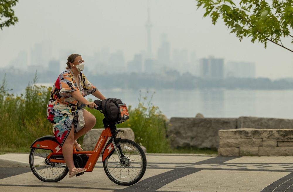 A masked cyclist rides along Humber Bay Shores Park as the smoky air partially obscures the Toronto skyline on Wednesday, July 15, 2026. 