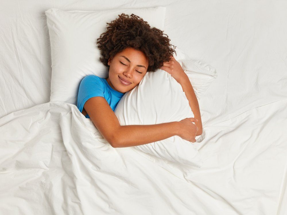 Satisfied pretty Afro American young woman has peaceful sleep, lies in bed on comfortable pillow, sees pleasant dreams, has gentle smile, enjoys softness of bedclothes, being under white blanket