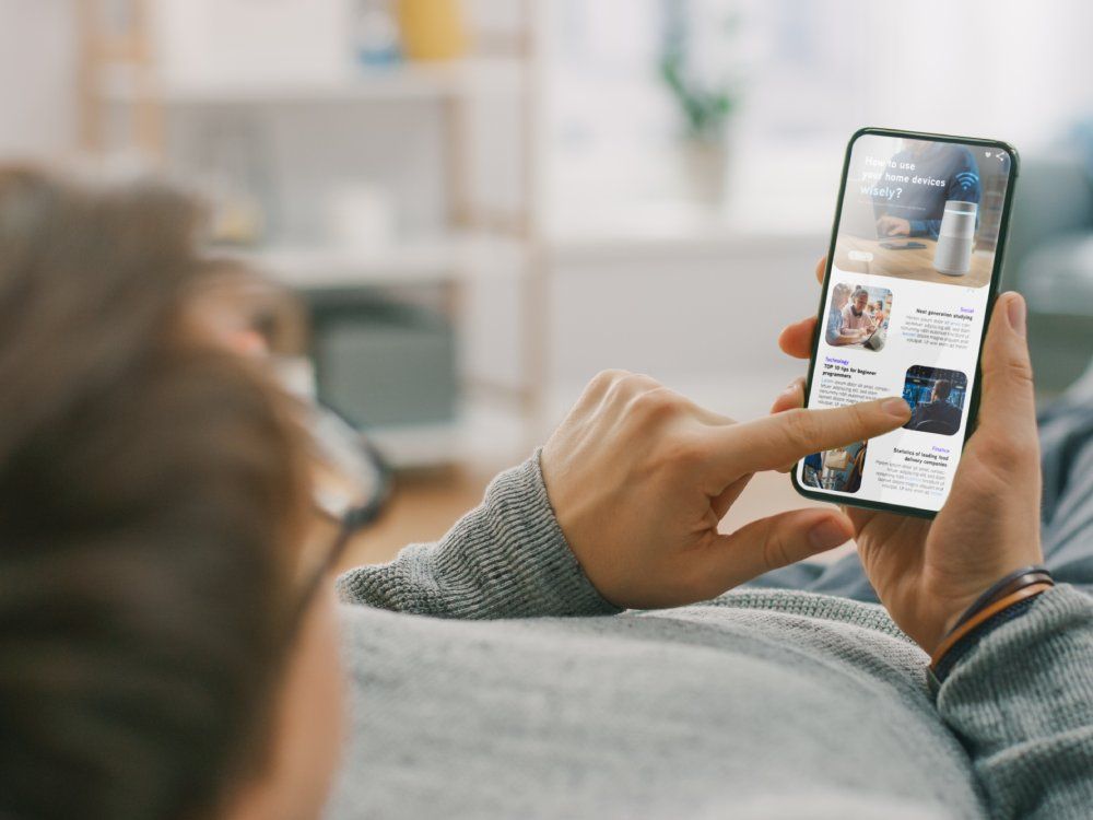 Young Man at Home is Lying on a Sofa and Using Smartphone for Scrolling and Reading News about Technological Breakthroughs. He's Sitting On a Couch in His Cozy Living Room. Over the Shoulder Shot