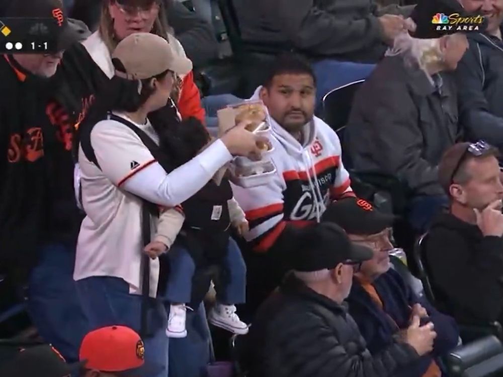 A mother returns to her seat while holding a baby and food during a San Francisco Giants game.