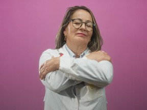 Woman doctor with grey hair and glasses hugging her shoulders with stethoscope in studio; self care serenity.