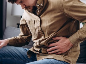 Upset male squeezing belly with hand because of abdominal pain. Unhappy young man sit on grey couch at home, suddenly feeling strong stomach ache, gastritis problem. Guy suffering from stomach ache