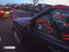 An SUV driver in Scotland, appears to be wearing headphones and looking at his cellphone, while a laptop rests on the console, near the gear shift. (Aberdeen Cycle Cam/YouTube screengrab)