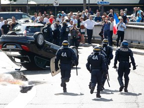 French riot police stand next to an overturned car as striking French taxi drivers demonstrate at the Porte Maillot to block the traffic on the Paris ring road during a national protest against car-sharing service Uber, in Paris, France, June 25, 2015. French taxi drivers stepped up protests against U.S. online cab service UberPOP on Thursday, blocking road access to airports and train stations in Paris and other cities. REUTERS/Charles Platiau