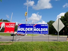 A pair of pedestrians walks past a collection of election signs for candidates in the London-West riding at the intersection of Riverside Drive and Sanatorium Road in London. Craig Glover/The London Free Press/Postmedia Network