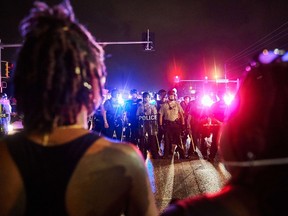 St. Louis County Police and Missouri State Highway Patrol troopers (C) stand guard as protesters (foreground) march on West Florissant Avenue in Ferguson, Missouri on August 9, 2015.  A day of peaceful remembrance marking the anniversary of 18-year-old black teen Michael Brown's killing by police in the US city of Ferguson came to a violent end on August 9 as gunfire left at least one protester injured.      AFP PHOTO / MICHAEL B. THOMAS
