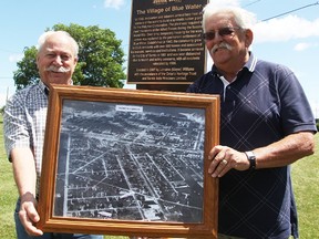 Gaston Croteau, left, and Serge Gauthier hold an aerial photo of the former Village of Blue Water in front of a historical plaque marking its spot in southern Sarnia. The former Blue Water residents are in a group organizing a 75th reunion commemorating the village's building in 1942. (Tyler Kula/Sarnia Observer/Postmedia Network)