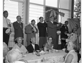 August 1, 1959. BC Premier WAC Bennett (playing violin) leads the Social Credit cabinet in a version of Happy Birthday to the Social Credit party at a Socred meeting in Kelowna. The politicians playing toy instruments are (L-R) provincial secretary Wes Black on toy drum, health minister Eric Martin on mouth organ, highways minister Flying Phil Gagliardi on accordion, attorney-general Robert Bonner on trumpet, Education minister Les Peterson on toy drum and Socred League president Noel Murphy on xylophone. Bennett's wife Mae is in front of him, with glasses. This was in the afternoon on the day Bennett shot a flaming arrow into a barge loaded with BC government bonds, a stunt that was known as "the burning of the bonds."