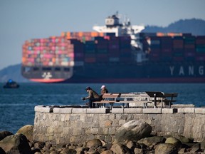 A man and woman read newspapers while sitting on the seawall as a container ship sits at anchor on English Bay, in Vancouver. Rail blockades across the country have led to an increase in the number of cargo ships waiting to load or unload according to the Port of Vancouver.