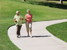 A senior couple takes a brisk walk in a park, holding dumbbells.