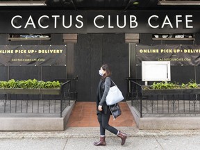 A woman wearing a mask walks past Cactus Club Cafe on Robson Street that has been boarded up after shifting to a delivery/takeout model only.
