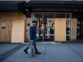 A man wearing latex gloves and a fabric mask on his face walks past a clothing store being boarded up, in Vancouver, on Thursday, April 16, 2020.