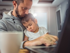 Father working on laptop at home office and holding son on his lap. Getty.