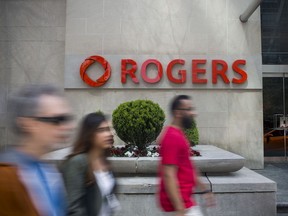 Pedestrians pass in front of Rogers Communications Inc. signage displayed outside a building in Toronto, Ontario, Canada, on Wednesday, May 17, 2017. Rogers Communications, Canada's largest wireless carrier, is leveraging organic growth in the country's wireless market to expand its subscriber base. Photographer: Brent Lewin/Bloomberg ORG XMIT: 700052488