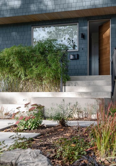 Concrete steps anchor the front entry, leading to a front porch that's partially sheltered by the home's second storey above. Simple perennial foliage and bamboo complement the modern minimalist design.