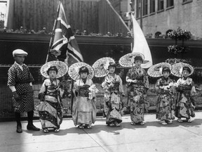 A line of young girls bedecked in kimono and parasols and a boy wearing a hapi and newsboy cap posing on a sidewalk in front of the firehall on Cordova Street in Vancouver in 1928. Between them and the building is a union jack and Japanese flag. Courtesy of Nikkei National Museum and Cultural Centre.