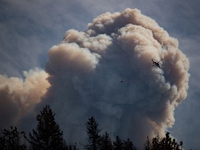A helicopter carrying a water bucket flies past a pyrocumulus cloud, also known as a fire cloud, produced by the Lytton Creek wildfire burning in the mountains above Lytton, B.C., on Sunday, August 15, 2021.