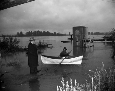1948 Fraser River Flood - Mission - A man in a suit stands in shallow flood waters while a nearby man watches from a canoe. 1948. Vancouver Sun.