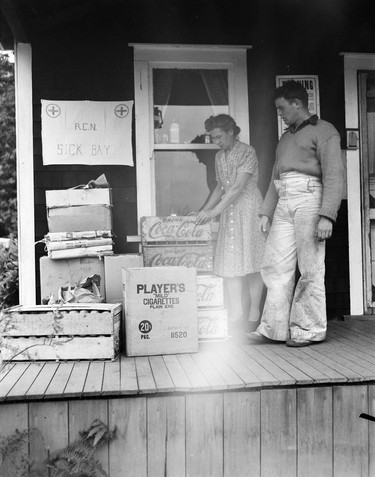 1948 Fraser River Flood - Nicomen Island & Deroche - A man and woman inspect boxes of cigarettes and other goods stacked on a porch and underneath a sign reading: 'R.C.N. Sick Bay.' 1948. Vancouver Sun.