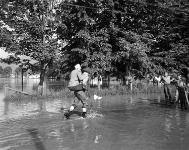 1948 Fraser River Flood - Rosedale - Children watch as a man carries a woman on piggyback through ankle-deep water. 1948. Vancouver Sun.