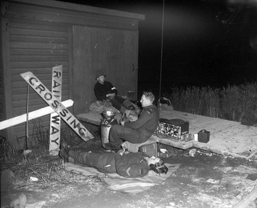 1948 Fraser River Flood - Sumas - Yarrows - Three men, at least two of whom are servicemen, rest and listen to a radio next to a downed railway crossing sign. Vancouver Sun. 1948.